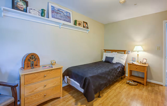 A small bedroom with a single bed covered in a dark quilt and white pillows. There is a wooden nightstand with a lamp, picture frames, and a pair of slippers on the floor beside it. On the opposite side, there is a wooden dresser with a vintage radio, a book, and a decorative item. Above the dresser, a white shelf holds framed pictures and decorative vases. The room has light-colored walls and wooden flooring.