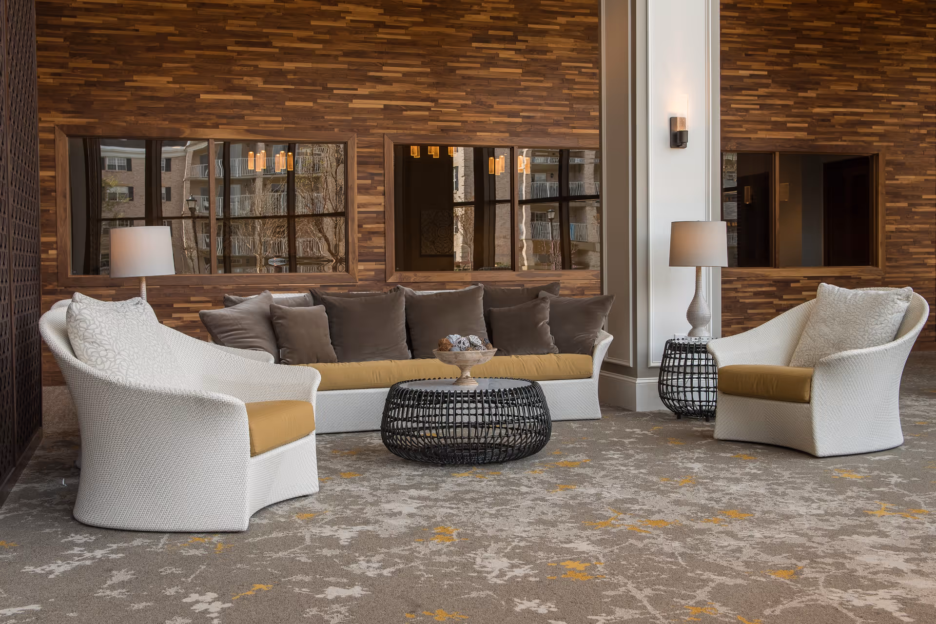 A modern lounge area with a white woven sofa and two matching armchairs with mustard cushions, a round black woven coffee table, two side tables with lamps, and a wooden accent wall with three rectangular windows reflecting the outside buildings.