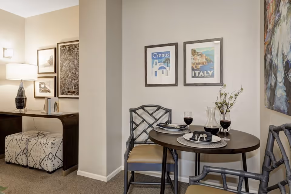 A cozy dining area with a small round table set for two with plates, silverware, and glasses of red wine. Two chairs with cushioned seats are placed around the table. On the wall above the table are two framed travel posters labeled Cyprus and Italy. To the left, there is a console table with a lamp, books, and decorative items, along with two patterned ottomans underneath. The walls are painted light beige, and the carpet is a neutral tone.