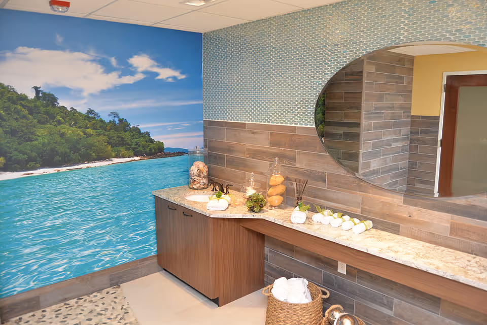 A bathroom with a large wall mural depicting a tropical beach scene with blue water and green trees. The bathroom features a long countertop with a sink, decorative jars, rolled towels, and a large oval mirror. The walls have a combination of blue mosaic tiles and wood-like tiles. A basket with rolled white towels is placed on the floor.