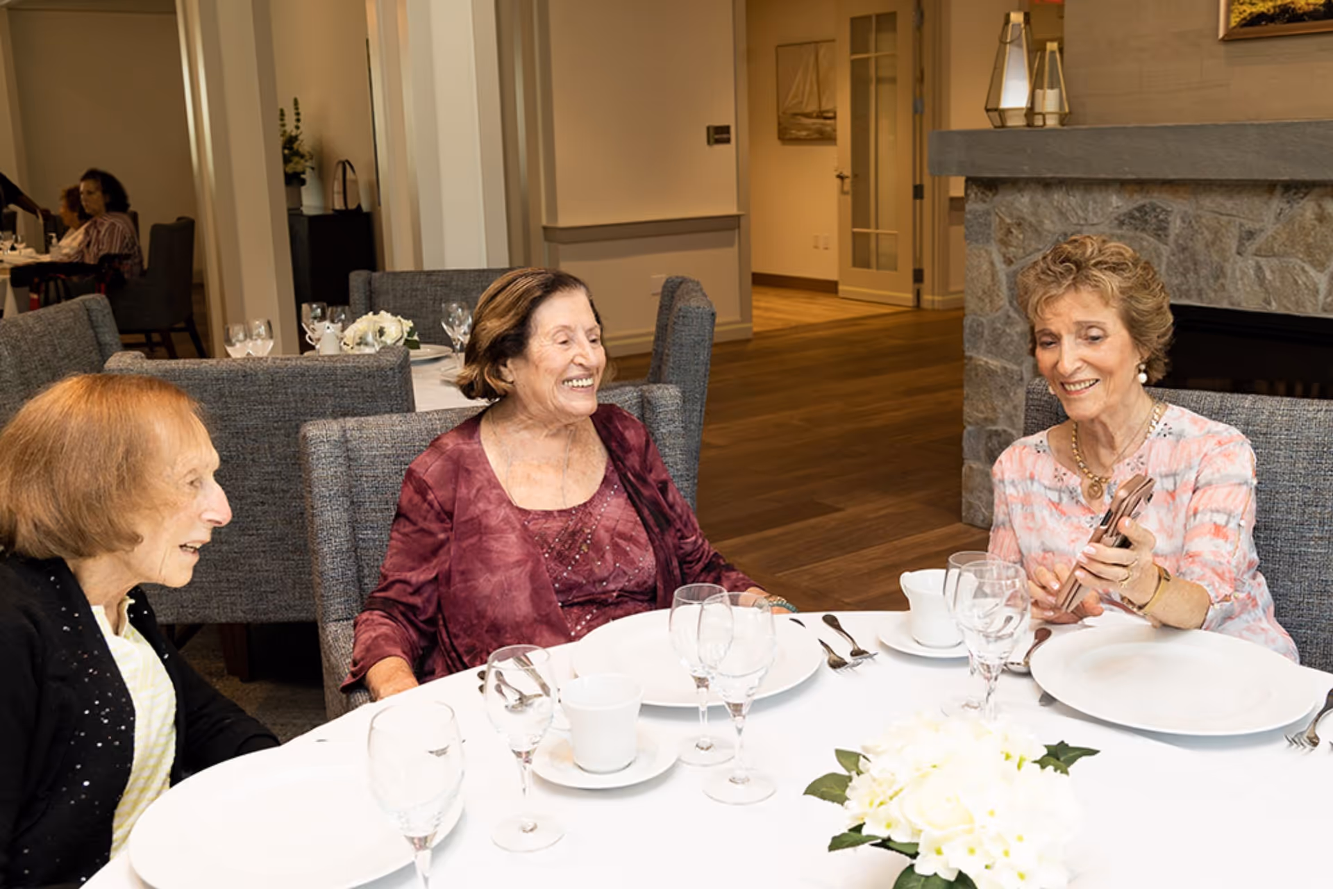 Three elderly women sitting around a dining table in a well-lit room, smiling and engaging with each other. The table is set with white plates, cups, and glassware, and there is a white floral centerpiece. The background shows more dining tables and a stone fireplace.