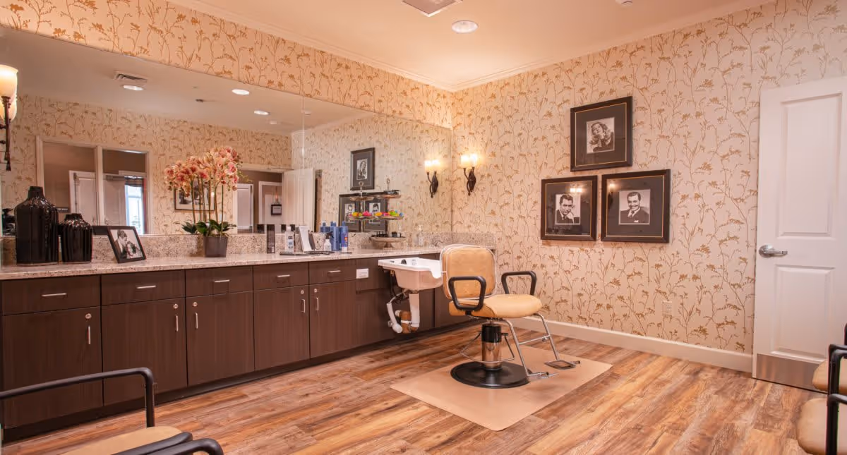 Interior of a salon room with a single salon chair in front of a large mirror. The room has wooden flooring, floral wallpaper, and a long countertop with various hair care products, a potted orchid, and framed photos. There are three framed black and white portraits on the wall and two wall-mounted light fixtures.