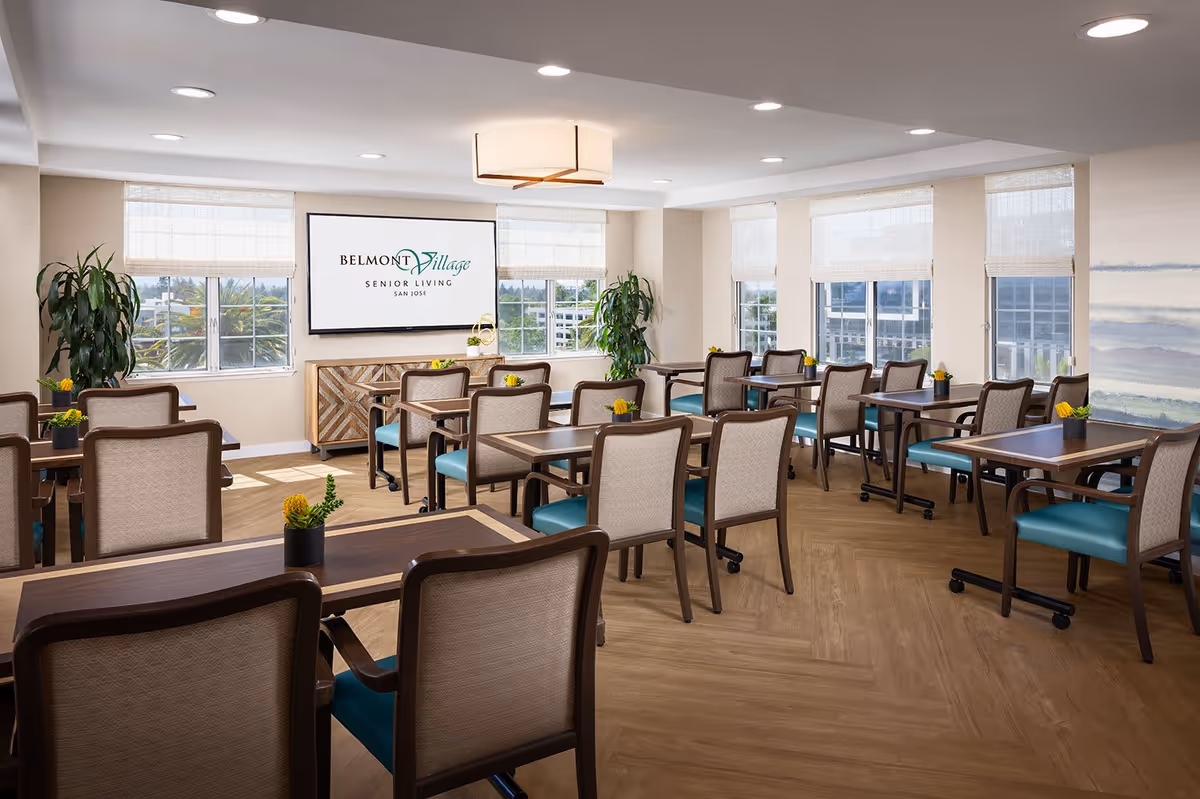 A bright and spacious dining room in Belmont Village Senior Living San Jose with multiple wooden tables and chairs arranged neatly. The room features large windows with white blinds allowing natural light to fill the space. There are small potted plants on each table and larger plants near the windows. A large screen on the wall displays the Belmont Village Senior Living San Jose logo.