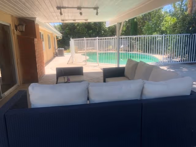 Covered outdoor patio area with cushioned seating and a view of a fenced swimming pool surrounded by trees and greenery.