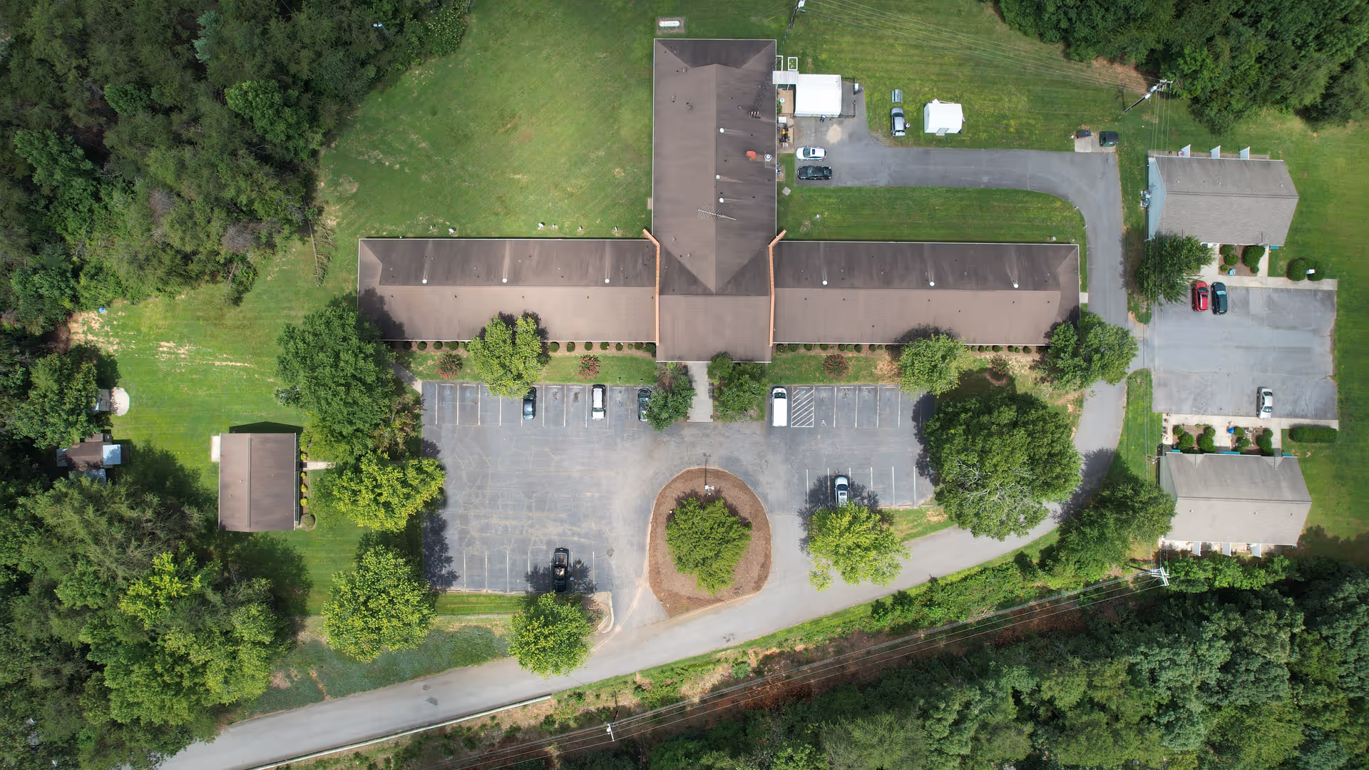Aerial view of Yadkin Valley Senior Living facility showing a large building with a T-shaped layout, surrounded by green lawns, trees, parking lots with several cars, and smaller adjacent buildings.
