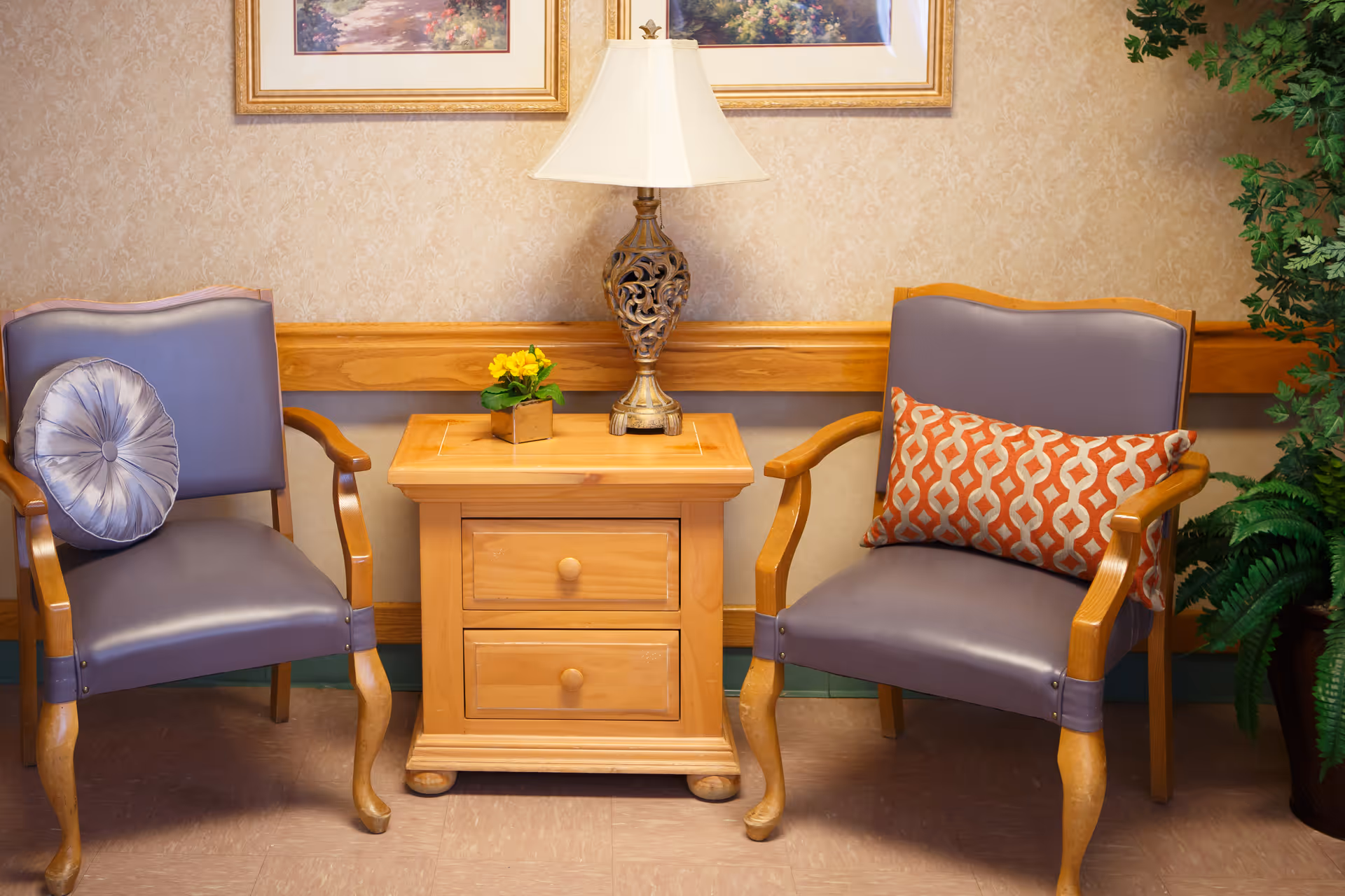 Two wooden armchairs with purple cushions and decorative pillows flank a wooden side table with two drawers. On the table is an ornate lamp and a small potted yellow flower. The background features beige patterned wallpaper, two framed landscape paintings, and a green potted plant on the right side.