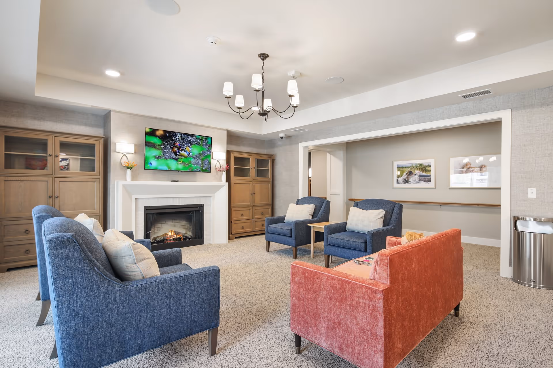 A cozy living room area with a red sofa and four blue armchairs arranged around a fireplace with a TV mounted above it. The room features beige carpet, two wooden cabinets flanking the fireplace, a chandelier, and framed pictures on the walls.