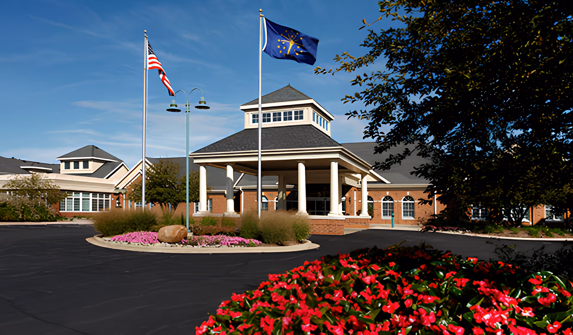 Exterior view of a senior living facility building with a covered entrance supported by white columns, two flagpoles displaying the American and Indiana state flags, landscaped flower beds with pink and red flowers, and a clear blue sky.
