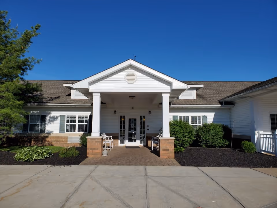 Front exterior view of The Residence of Chardon building with a covered entrance supported by white columns, benches on either side, surrounded by green shrubs and plants under a clear blue sky.