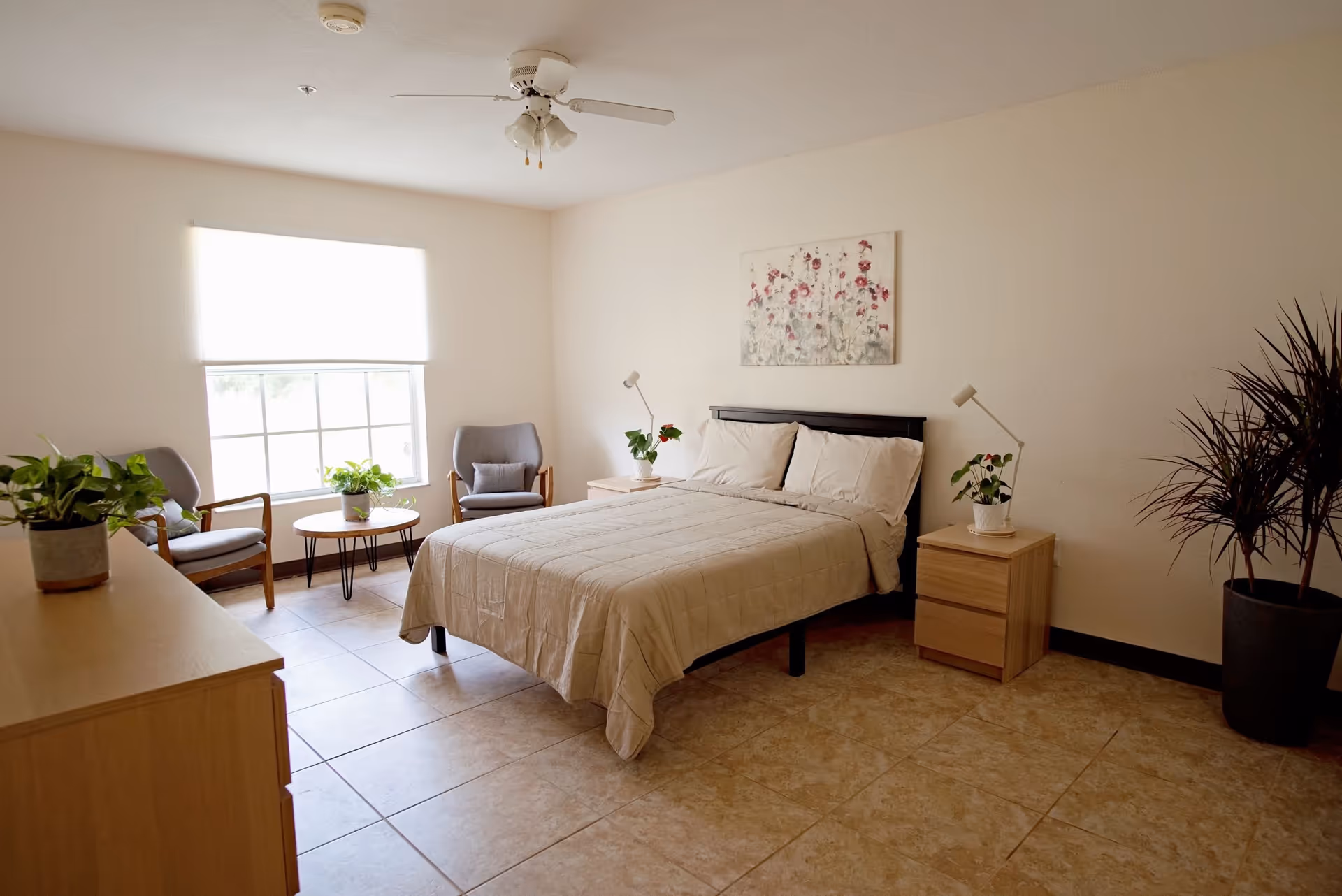 A bright and clean bedroom with a double bed covered in beige bedding, two wooden nightstands with small lamps and potted plants, a dresser with a plant, two gray armchairs with a small round table between them near a large window with a white blind, a ceiling fan, and a large potted plant in the corner.