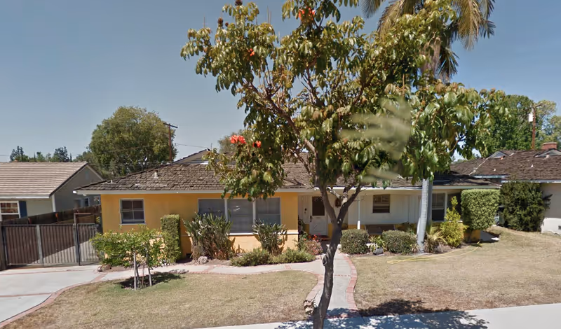 Single-story residential building with a yellow exterior and a shingled roof, surrounded by a lawn with a tree and various shrubs in front. A paved walkway leads to the front door, and neighboring houses are visible on either side under a clear blue sky.