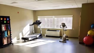 A senior living facility exercise room with a treadmill, stationary bike, and a rack holding various exercise balls and weights. Large windows allow natural light to fill the room.