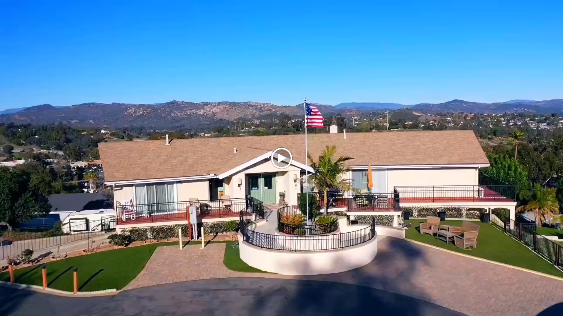 A single-story building with a tan roof and light-colored walls, featuring a central entrance with green double doors. In front of the building is a circular raised planter with an American flag and palm trees. There is a paved driveway and green lawn areas with outdoor seating. The background shows a mountainous landscape under a clear blue sky.