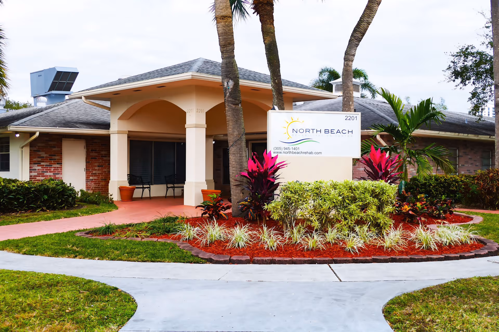 Exterior view of North Beach Rehabilitation Center building with a covered entrance, palm trees, and landscaped garden with colorful plants and shrubs. A sign in front displays the facility name, phone number, and website.