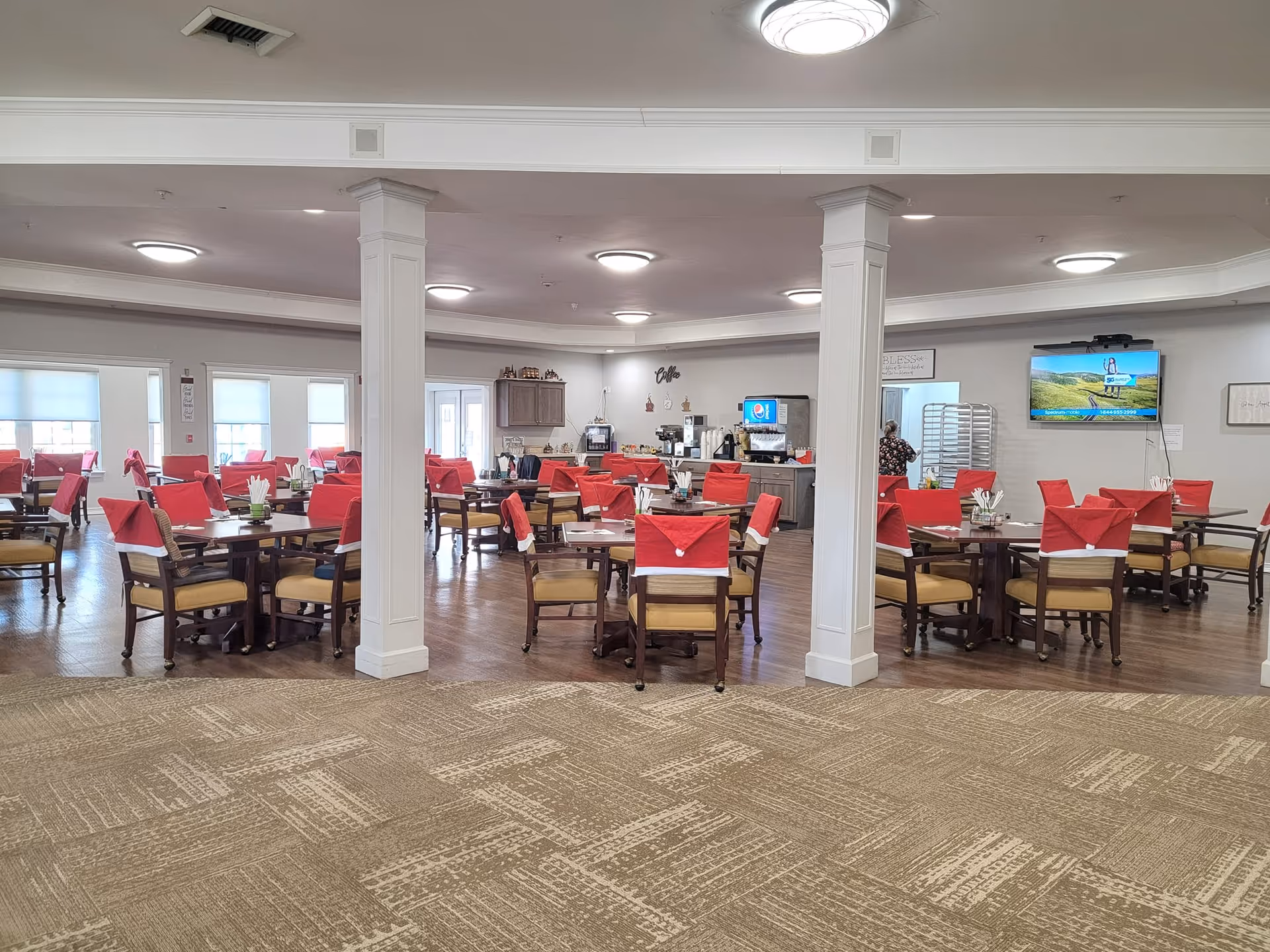 A spacious dining room in Colonial Assisted Living & Independent Living with multiple wooden tables and chairs. Each chair is decorated with a red and white Santa hat cover. The room has beige carpet and wood flooring, white walls, and ceiling lights. There is a coffee station and a soda machine against the far wall, and a television mounted on the right wall. A person is seen near the back of the room.