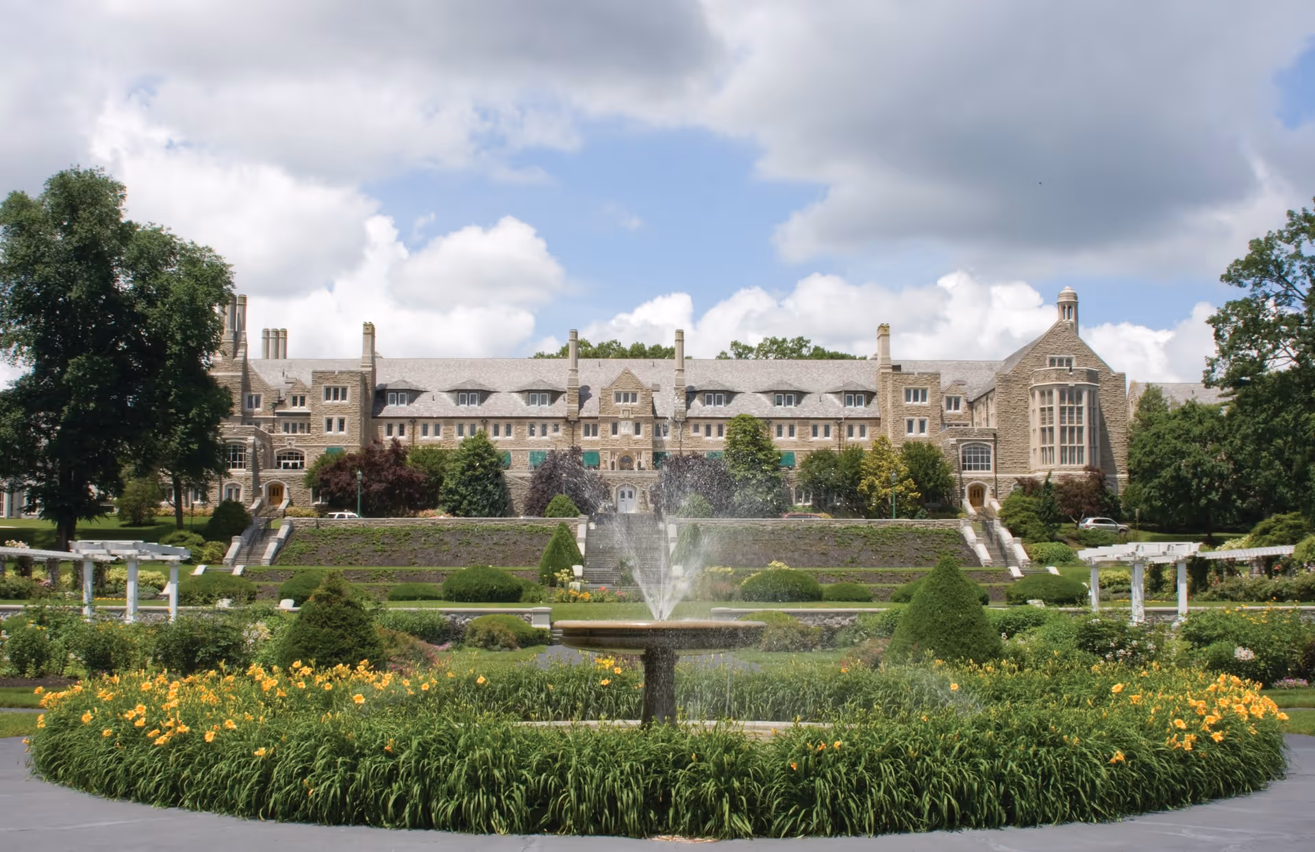A large stone building with multiple windows and chimneys, set behind a landscaped garden with a central fountain surrounded by yellow flowers and greenery under a partly cloudy sky.
