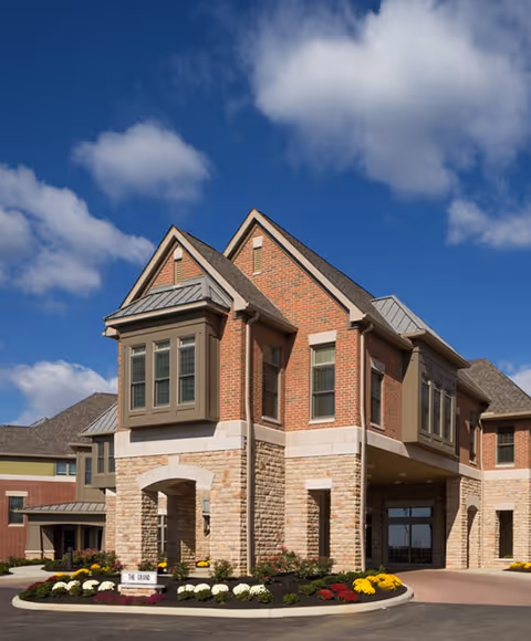 Exterior view of a senior living facility building with a combination of brick and stone facade under a blue sky with some clouds. The building has multiple windows and a covered entrance. There are landscaped flower beds with colorful flowers near the entrance.