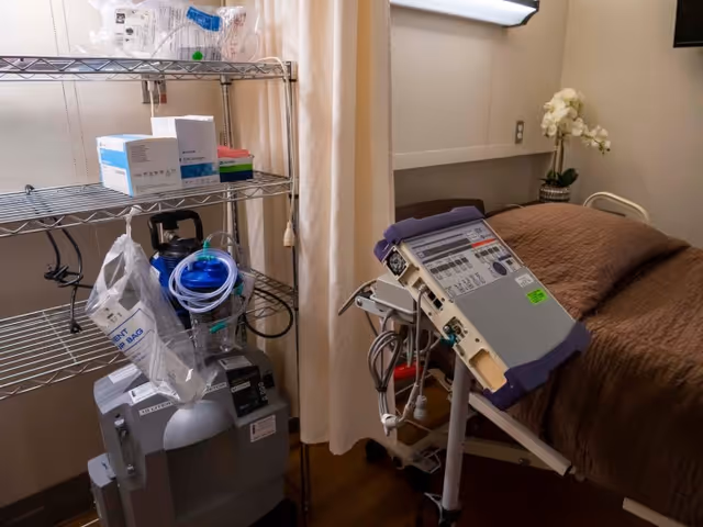A medical room with a hospital bed covered in a brown blanket, a medical device on a stand next to the bed, and a metal shelving unit holding medical supplies and equipment. A beige curtain is partially drawn in the background, and a white flower arrangement is on a shelf above the bed.