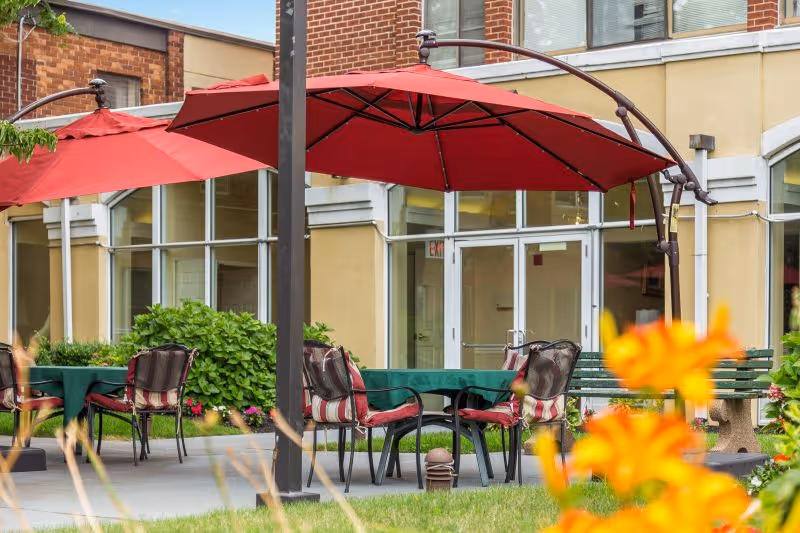 Outdoor seating area at Castle Senior Living with tables covered in green tablecloths, chairs with red and white striped cushions, and large red umbrellas providing shade. The background shows the building's exterior with large windows and a door.