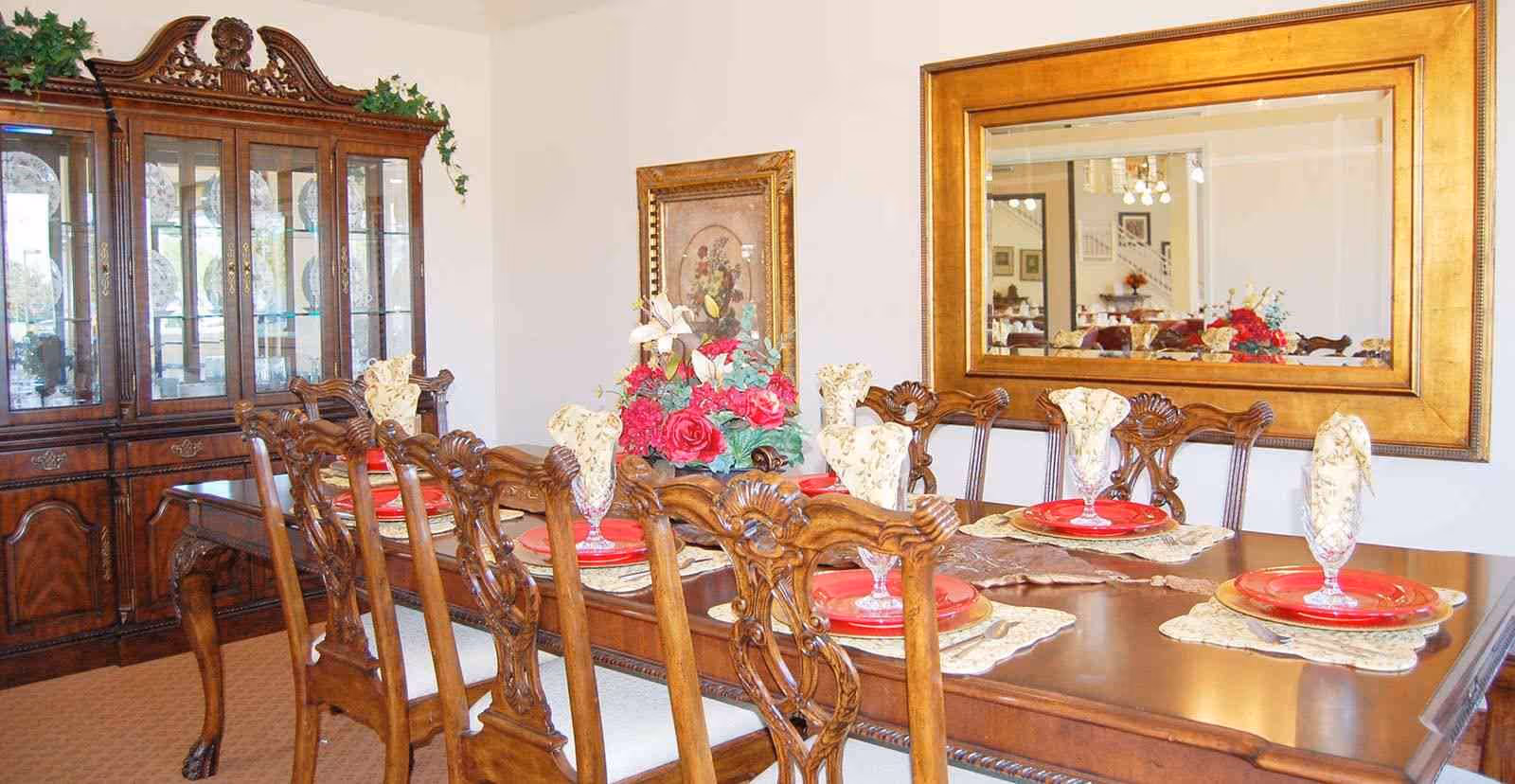 A formal dining room with a long wooden table set with red plates, glass goblets, and cream-colored napkins. The room features ornate wooden chairs, a large wooden china cabinet displaying plates, a floral centerpiece on the table, a framed painting on the wall, and a large gold-framed mirror reflecting part of the room.