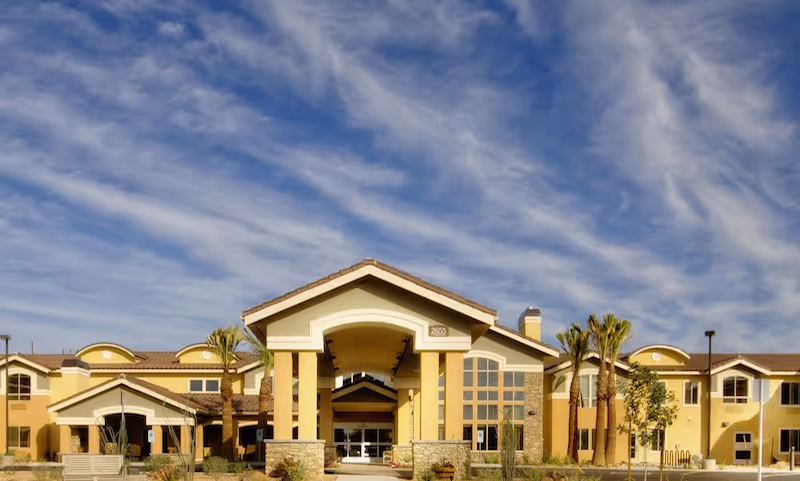 Front exterior view of The Groves Assisted Living & Memory Care building with a covered entrance, palm trees, and a partly cloudy sky above.
