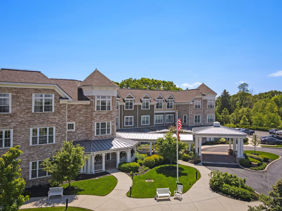 Exterior view of a senior living facility building with stone and siding facade, multiple windows, a covered entrance, a flagpole with an American flag, benches, trees, and a parking area under a clear blue sky.