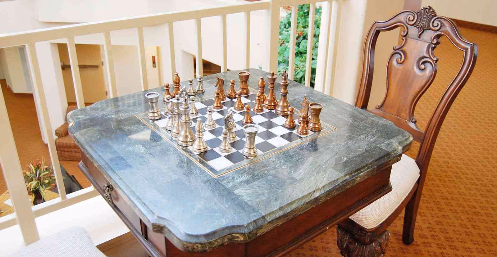 A marble-topped table with an inlaid chessboard and metal chess pieces set up for a game, accompanied by a carved wooden chair with a cushioned seat, situated indoors near a railing and overlooking a lower level with plants.