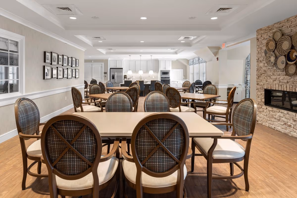 A spacious dining room with multiple tables and cushioned chairs arranged neatly. The room features a modern kitchen area in the background with white cabinets, stainless steel appliances, and pendant lighting. The walls are decorated with framed pictures and a brick accent wall with decorative woven baskets. The floor is wooden, and the room is well-lit with recessed ceiling lights.