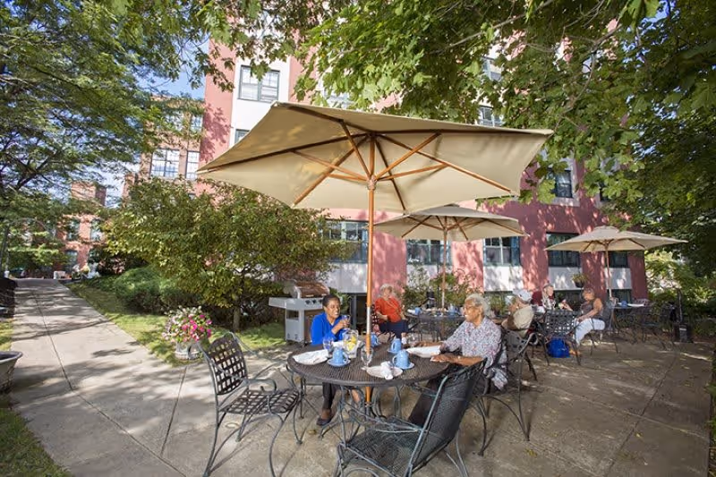Outdoor patio area at Standish Village Assisted Living & Compass Memory Support Community with several elderly people sitting at round metal tables under large beige umbrellas, surrounded by trees and greenery, adjacent to a multi-story building.