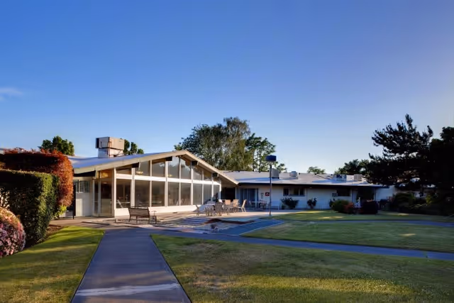 Exterior view of a single-story building with large windows and a slanted roof, surrounded by well-maintained green lawns, bushes, and trees under a clear blue sky. There are outdoor benches and tables near the building entrance.
