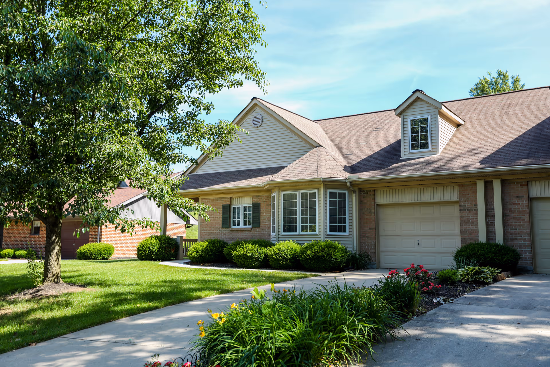 Exterior view of a single-story residential building with a garage, surrounded by green grass, bushes, and a tree under a clear blue sky.