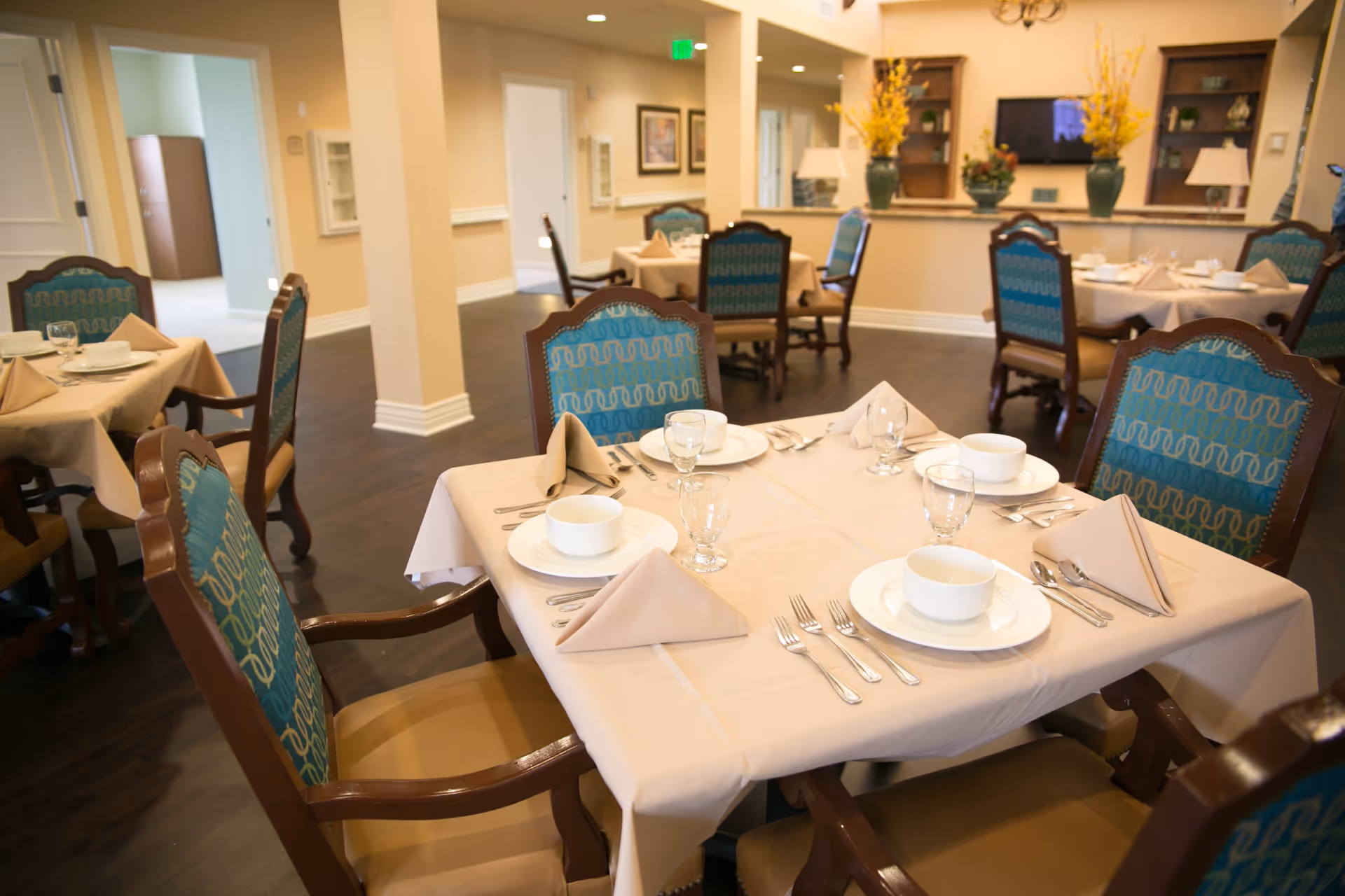 A dining room in Buckner Villas with tables set for a meal. Each table has beige tablecloths, neatly folded beige napkins, white plates, bowls, glasses, and silverware. The chairs have wooden frames with blue patterned upholstery. The room has warm lighting, wooden flooring, and decorative plants on a counter in the background.
