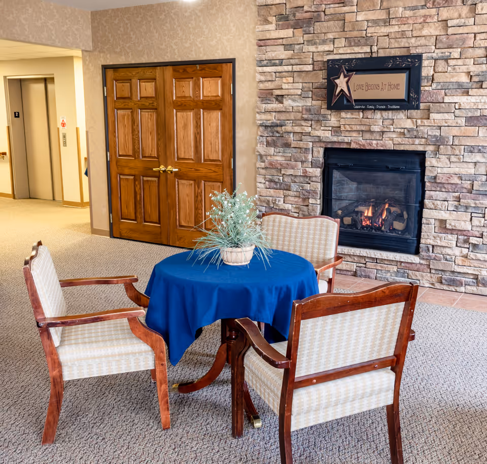A cozy seating area in a senior living facility featuring a round table covered with a blue tablecloth, surrounded by four wooden chairs with cushioned seats and backs. A potted plant is placed on the table. Behind the seating area is a stone fireplace with a fire burning and a decorative sign above it that reads 'Love Begins At Home.' To the left, there are double wooden doors and an elevator with the number 2 displayed.