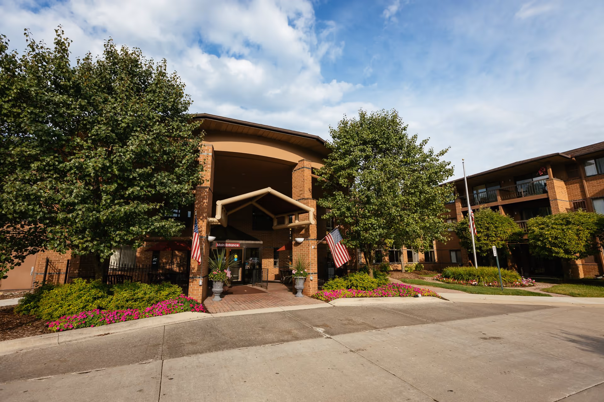 Exterior view of a senior living facility named Bellbrook showing the main entrance with a covered walkway, two American flags, well-maintained landscaping with trees and colorful flowers, and a clear blue sky with some clouds.