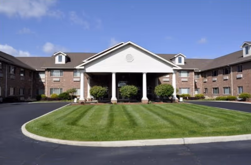 Front exterior view of a senior living facility with a large covered entrance supported by white columns, surrounded by well-maintained green lawn and bushes, under a clear blue sky.