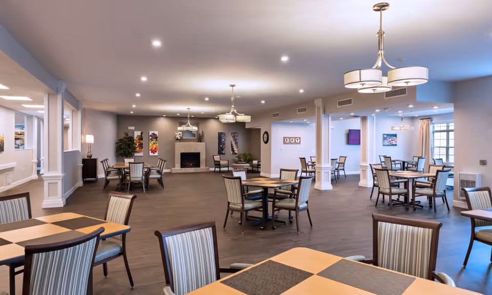 Spacious dining room with multiple tables and chairs arranged neatly. The room features a modern fireplace on the far wall, decorative artwork, and soft lighting from ceiling fixtures. The floor is wooden, and the walls are painted in neutral tones, creating a warm and inviting atmosphere.