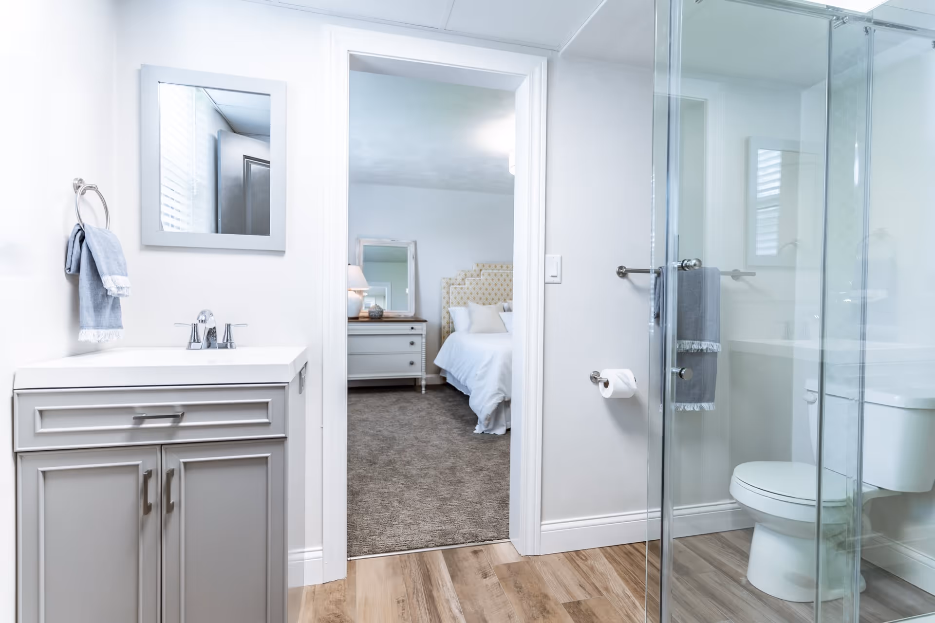 A bright bathroom with a gray vanity and sink on the left, a towel hanging on a ring, a glass-enclosed shower on the right, and a toilet next to it. Through the open doorway, a bedroom with a bed, nightstand, lamp, and mirror is visible.