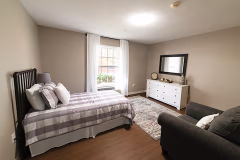 A cozy bedroom with a single bed covered in a checkered purple and white bedspread, a gray armchair with a white pillow, a white dresser with decorative items and a large mirror above it, a window with white curtains letting in natural light, and a patterned area rug on a wooden floor.