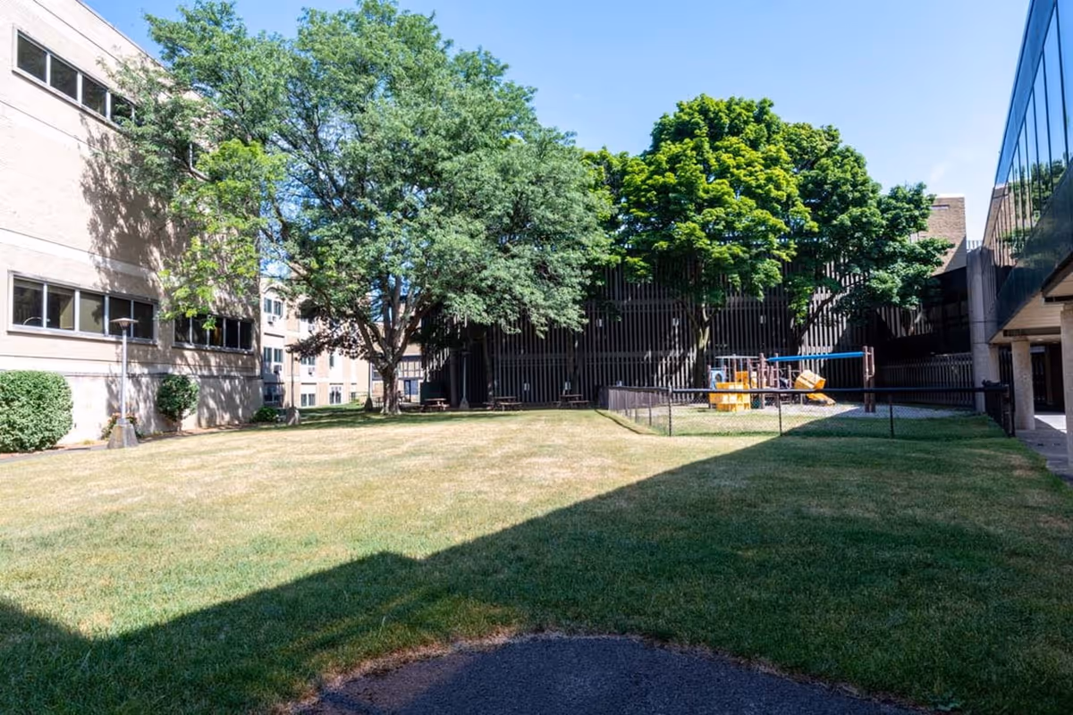 Outdoor grassy courtyard area at RRH Unity Living Center with several large trees providing shade, a small fenced playground with slides and climbing equipment, and surrounding multi-story buildings with windows.