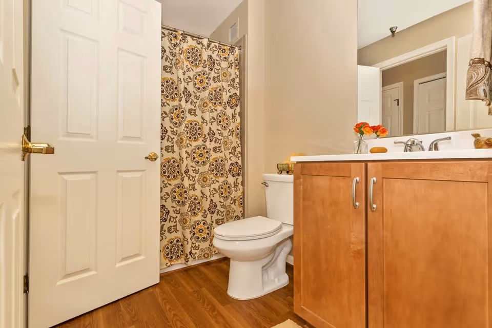 A bathroom with a white toilet, wooden floor, and a wooden vanity with a white countertop. There is a large mirror above the vanity, a floral-patterned shower curtain, and a partially open white door. A small vase with orange flowers and a soap dish are on the countertop.