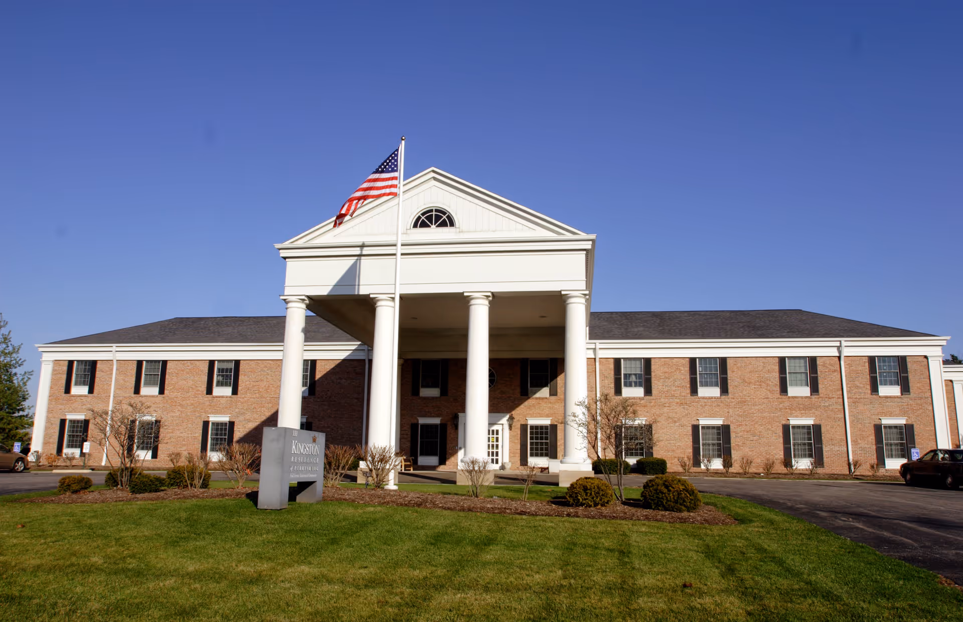 Front exterior view of Kingston Residence of Perrysburg, a two-story brick building with white columns and an American flag flying on a flagpole in front. There is a sign on the lawn with the facility's name and a clear blue sky above.