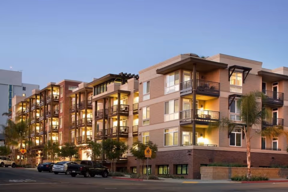 Exterior view of a modern multi-story residential building with balconies and large windows, surrounded by palm trees and parked cars along the street during dusk.