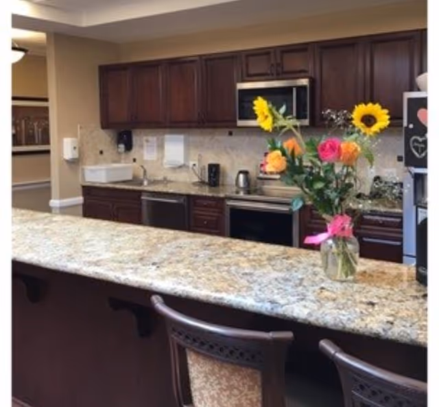 A kitchen area with dark wooden cabinets, a granite countertop island with two chairs, a microwave, stove, dishwasher, and a vase with colorful flowers on the countertop.