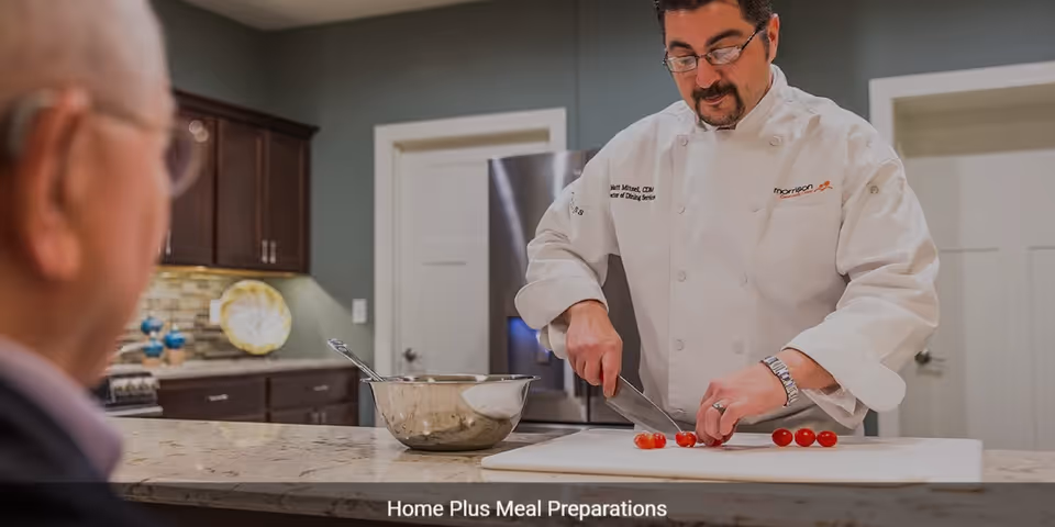 A chef wearing a white uniform is slicing cherry tomatoes on a cutting board in a kitchen. An elderly man is watching from the other side of the counter. The kitchen has dark wooden cabinets and a stainless steel refrigerator.