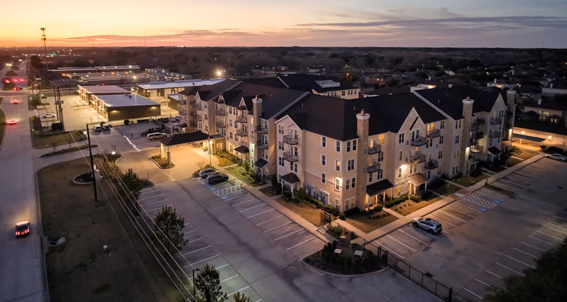 Aerial view of Park Creek Active Retirement facility at dusk, showing a large multi-story residential building with balconies, surrounded by parking lots and nearby commercial buildings under a colorful sunset sky.