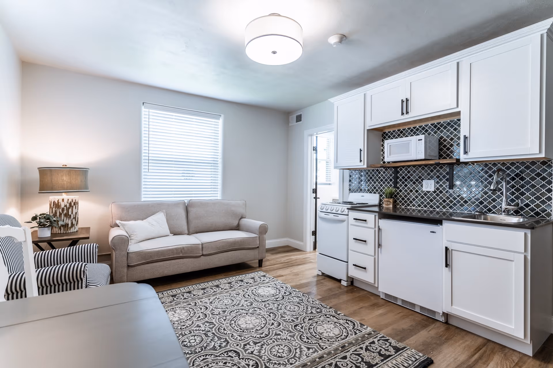 A bright and clean living area with a beige sofa, a striped armchair, a side table with a lamp and plant, and a kitchen area featuring white cabinets, a stove, microwave, sink, and a patterned tile backsplash. A patterned rug covers part of the wooden floor.