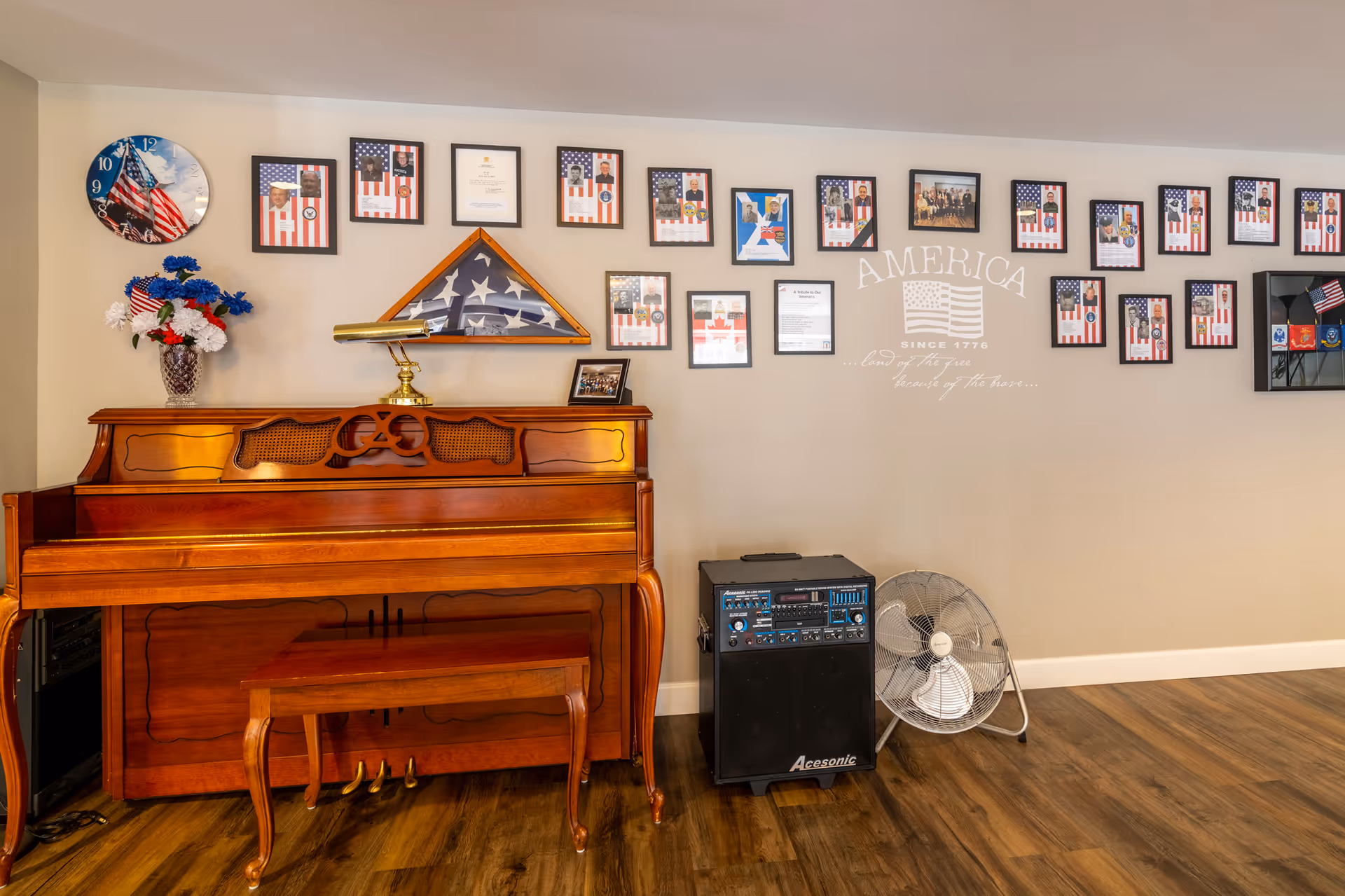 Interior room with a wooden upright piano and matching bench on a wood floor. Above the piano is a wall clock with an American flag design, a vase with red, white, and blue flowers, and a folded American flag in a triangular display case. The wall behind the piano is decorated with framed photos and certificates, many featuring American flags. There is also a speaker and a standing fan on the floor next to the piano. The wall has the word 'AMERICA' and a flag graphic with the text 'SINCE 1776 ...land of the free home of the brave...'.