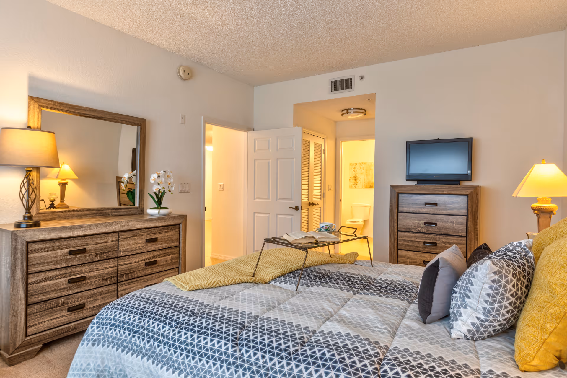 A cozy bedroom with a bed covered in a patterned gray and white comforter and multiple pillows. A wooden dresser with a large mirror and a lamp is on the left side of the room. A small TV sits on a wooden chest of drawers on the right side. The door to a bathroom is open, showing a toilet inside. The room is warmly lit with lamps on both sides.