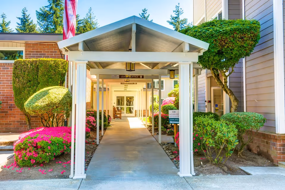 Covered walkway entrance to a building at Sagebrook Senior Living at Bellevue, surrounded by neatly trimmed bushes and colorful flowers, with an American flag visible on the left side.