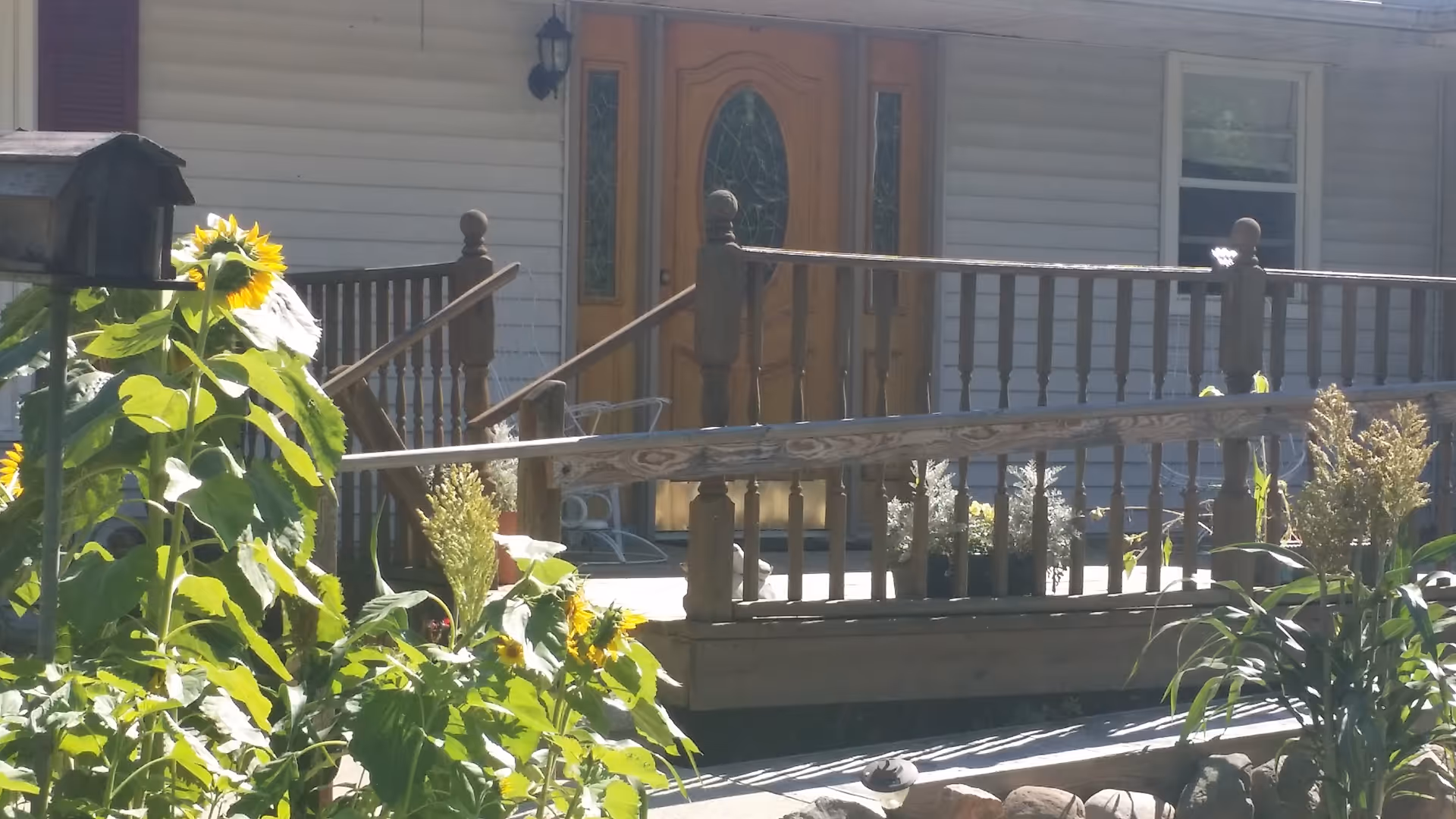 Front porch of a building with wooden railings and a wooden door with decorative glass panels. There are sunflowers and other plants in the foreground, and a mailbox is visible on the left side.